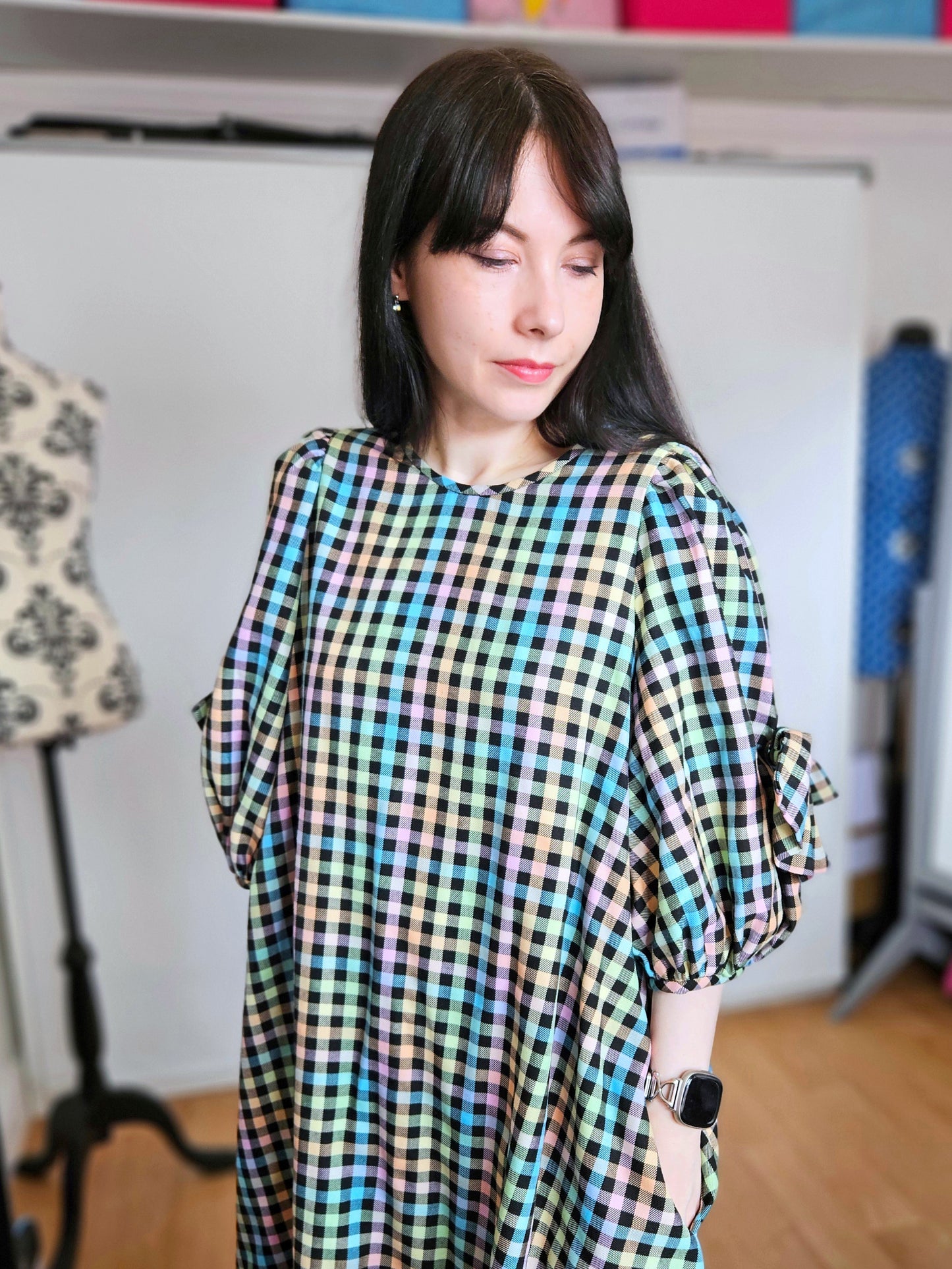 A woman wearing a colourful plaid dress with puff sleeves, standing in a room with a sewing mannequin and shelves in the background.