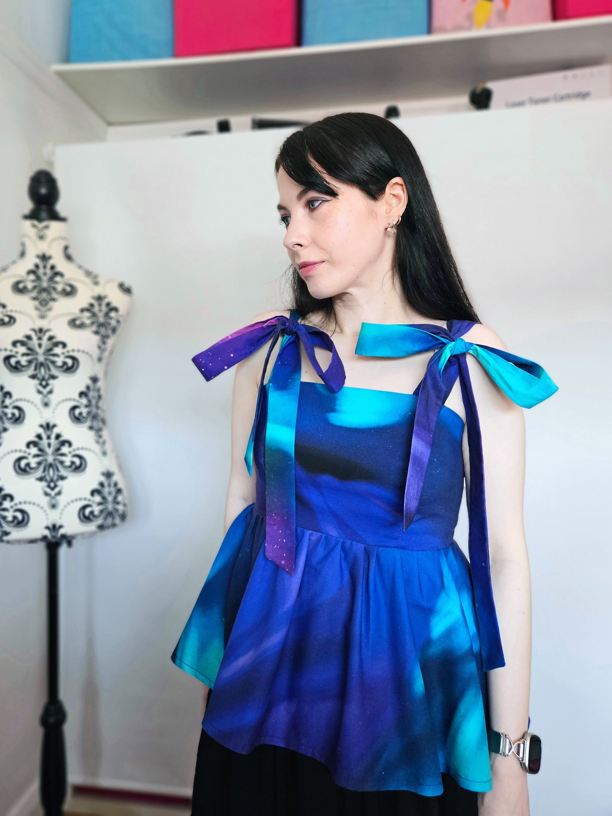 A woman wearing a sleeveless peplum top with a blue, purple, and teal Northern Lights pattern, featuring shoulder tie details with large bows, paired with a black skirt. She is standing in front of a white wall next to a floral patterned dress form.
