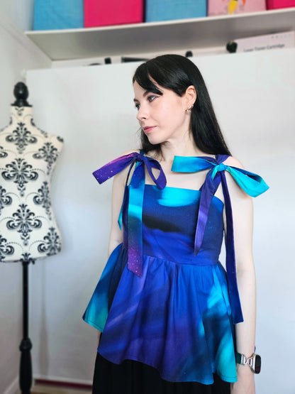 A woman wearing a sleeveless peplum top with a blue, purple, and teal Northern Lights pattern, featuring shoulder tie details with large bows, paired with a black skirt. She is standing in front of a white wall next to a floral patterned dress form.