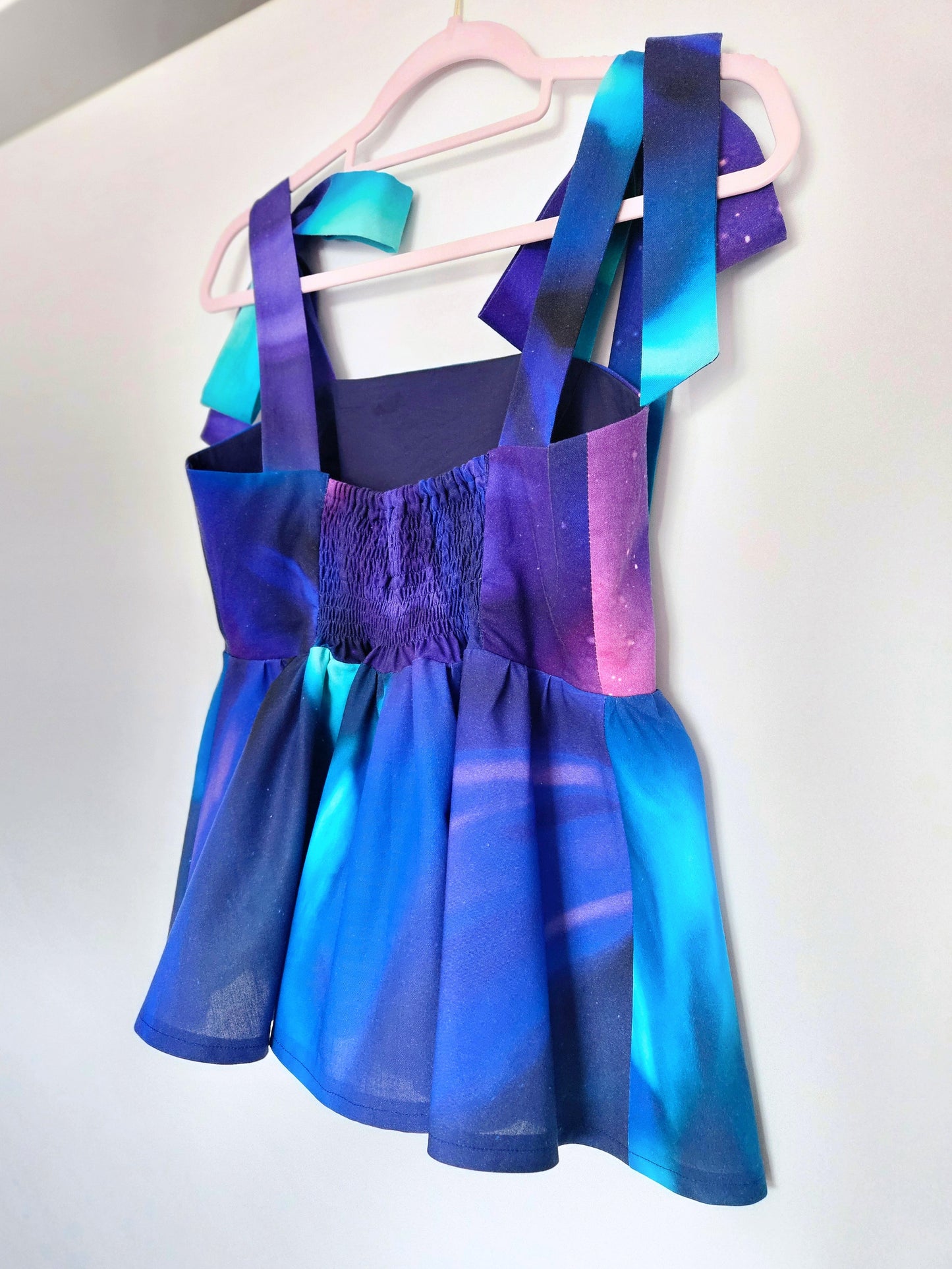 The back of a sleeveless peplum top with a blue, purple, pink and teal Northern Lights pattern, with large adjustable shoulder ties with large bows is hanging on a pale pink hanger against a white wall. The back has an elasticated, shirred panel.