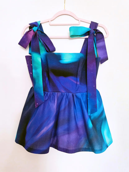 A sleeveless peplum top with a blue, purple, and teal Northern Lights pattern, with large adjustable shoulder ties with large bows is hanging on a pale pink hanger against a white wall.