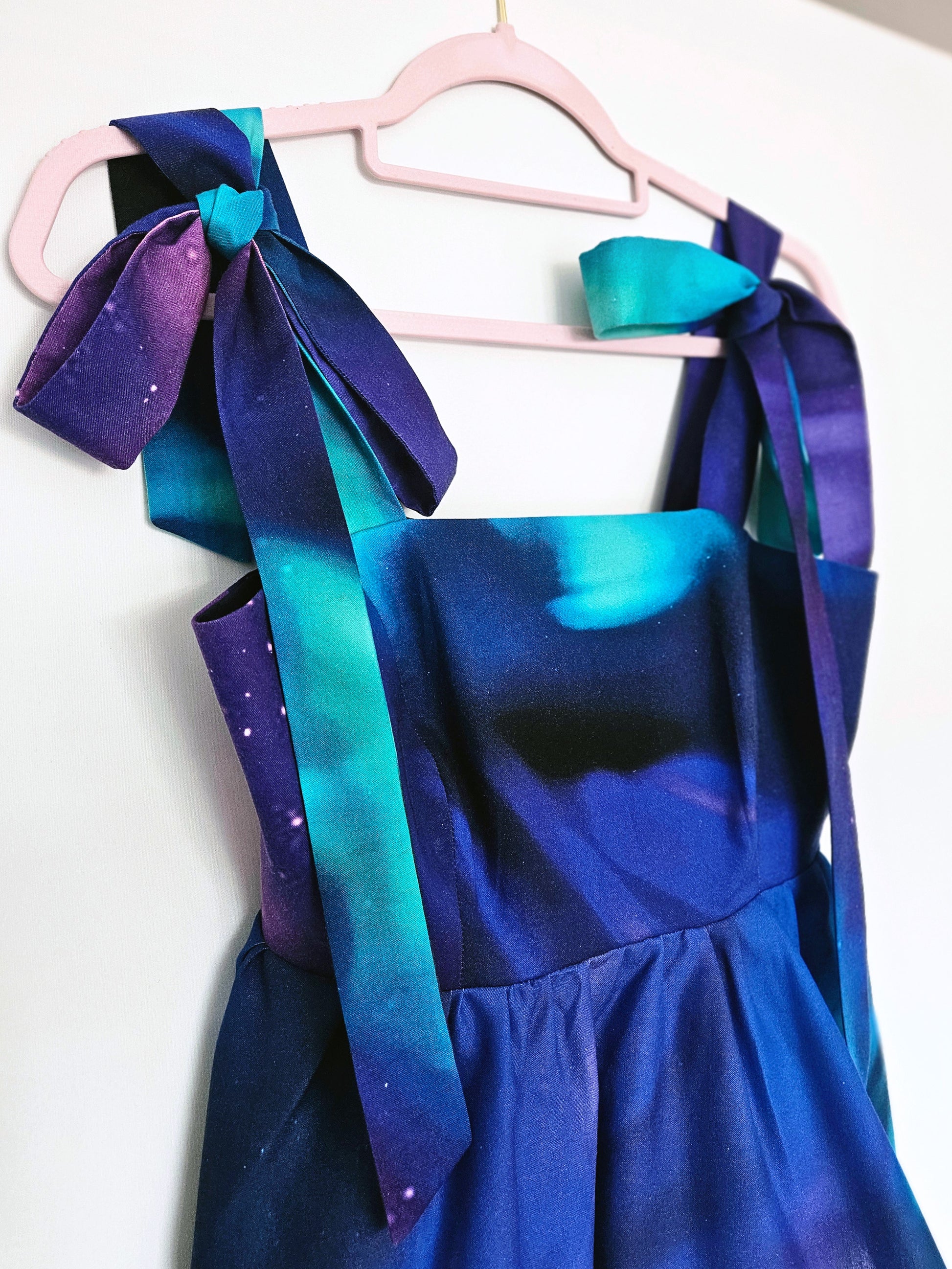 A blue, turquoise and purple Northern Lights print top hanging on a light pink hanger against a plain white wall. The top features wide shoulder straps with large bows and a gathered peplum.