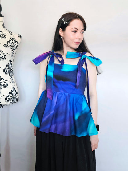 A woman wearing a sleeveless peplum top with a blue, purple, and teal Northern Lights pattern, featuring shoulder tie details with large bows, paired with a black skirt. She is standing in front of a white wall next to a floral patterned dress form.