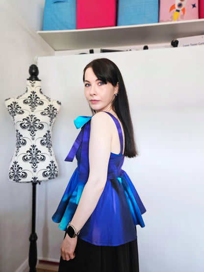 A woman wearing a sleeveless peplum top with a blue, purple, and teal Northern Lights pattern, featuring shoulder tie details with large bows, paired with a black skirt. She is standing in front of a white wall next to a floral patterned dress form.