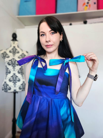 A woman wearing a sleeveless peplum top with a blue, purple, and teal Northern Lights pattern, featuring shoulder tie details with large bows. She is holding one of the shoulder bows while standing in front of a white wall next to a floral patterned dress form.