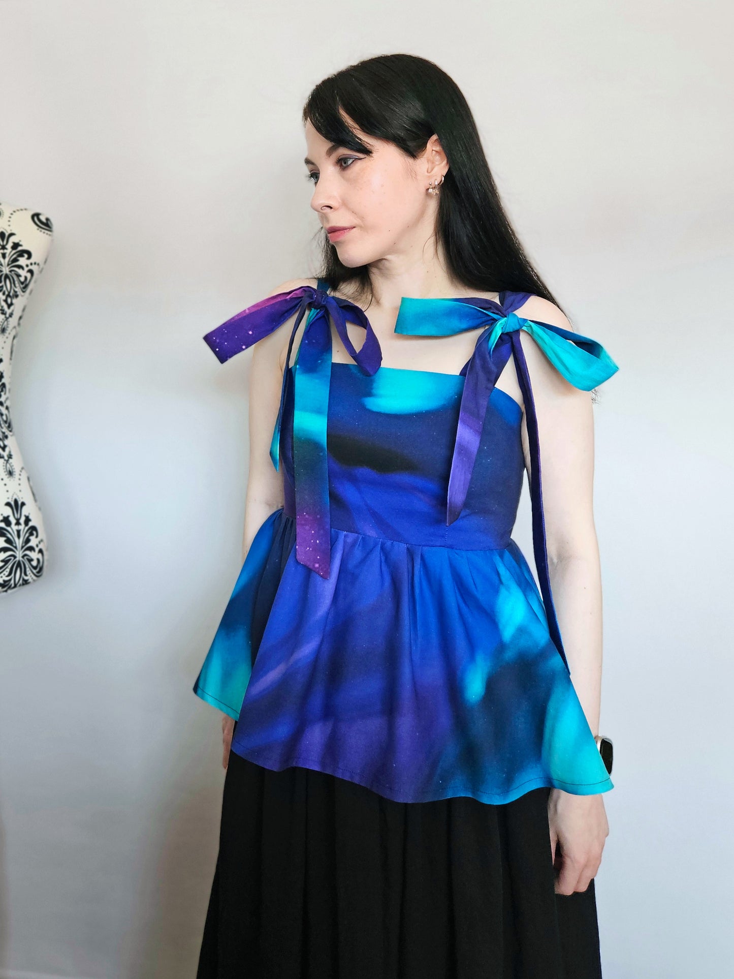 A woman wearing a sleeveless peplum top with a blue, purple, and teal Northern Lights pattern, featuring shoulder tie details with large bows, paired with a black skirt. She is standing in front of a white wall next to a floral patterned dress form.