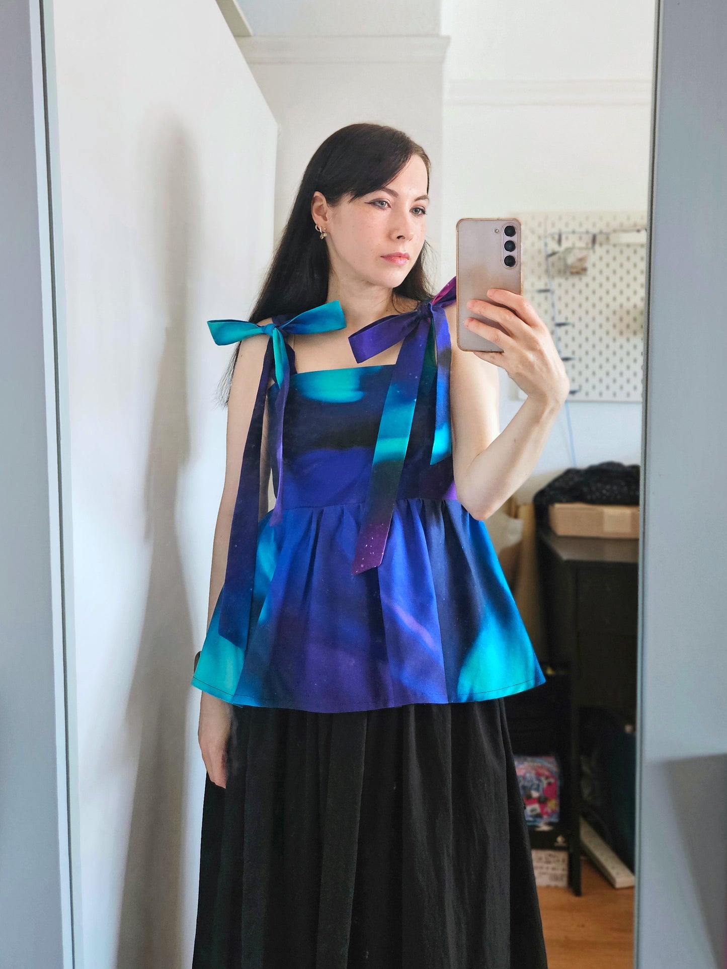 A woman wearing a sleeveless peplum top with a blue, purple, and teal Northern Lights pattern is taking a mirror selfie. The top has shoulder tie details with large statement bows, paired with a black skirt.