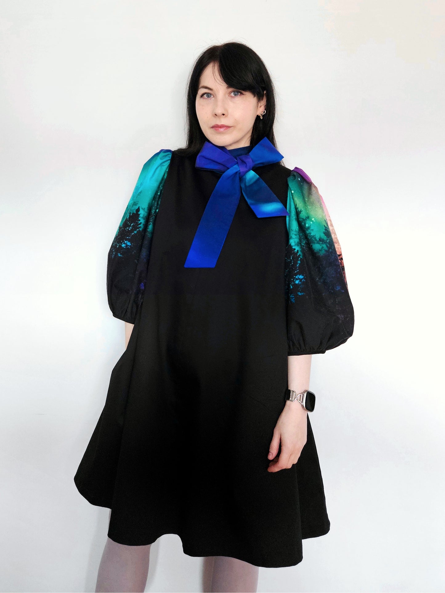 Woman wearing a black dress with colourful aurora print sleeves and a blue bow against a white background