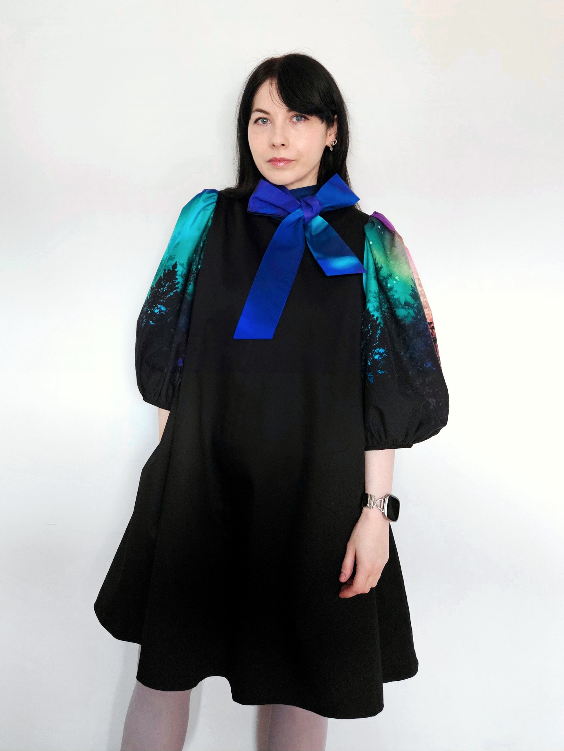 Woman wearing a black dress with colourful aurora print sleeves and a blue bow against a white background