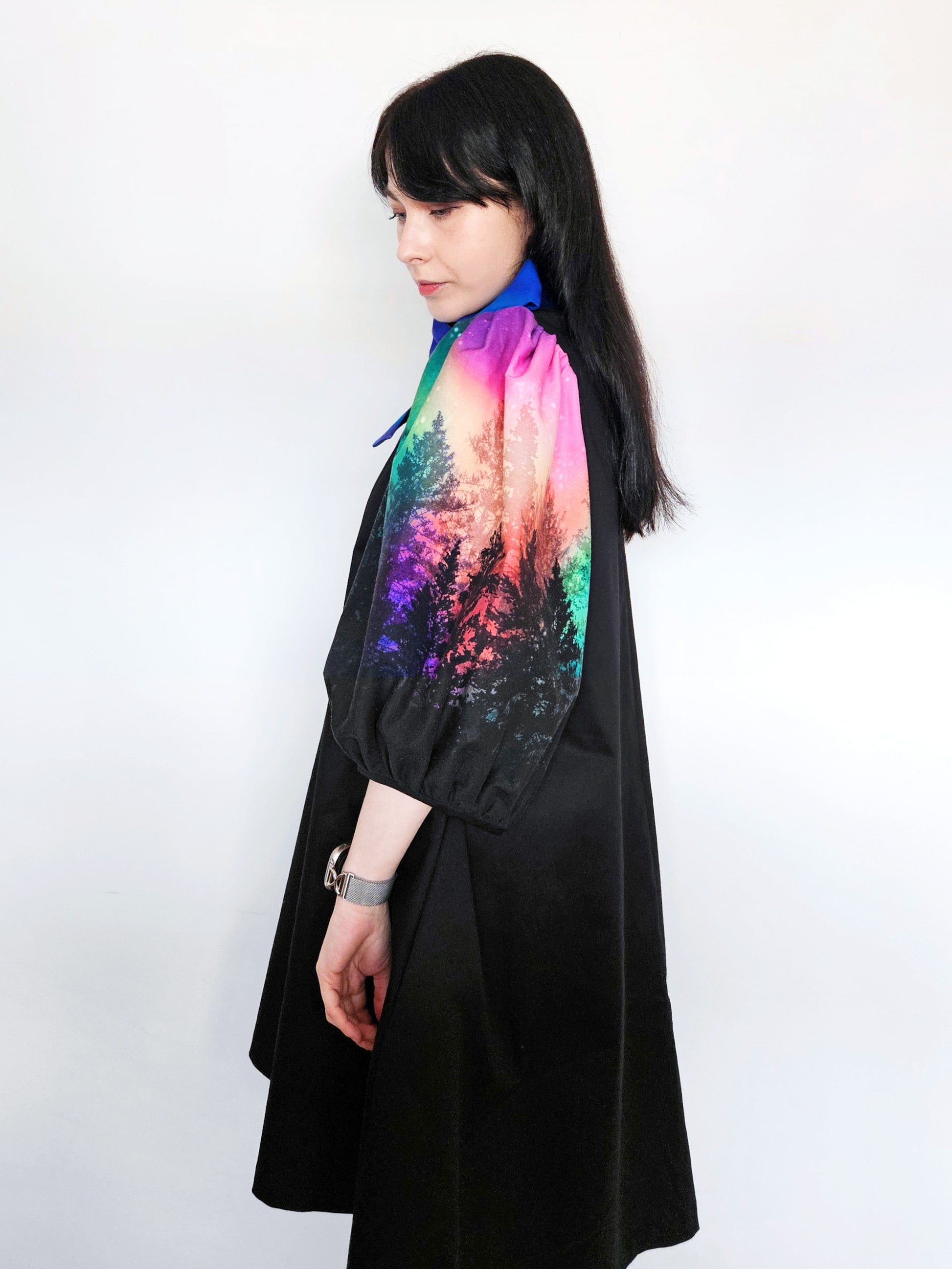 Woman wearing a black dress with colourful aurora print sleeves and a blue bow against a white background