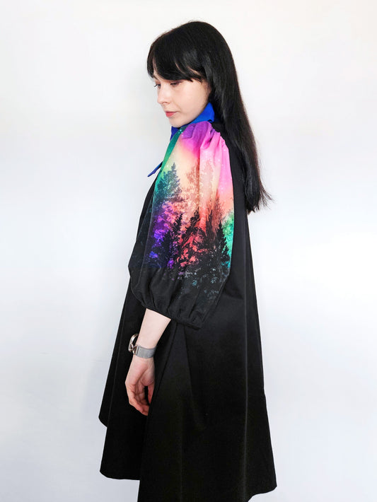 Woman wearing a black dress with colourful aurora print sleeves and a blue bow against a white background