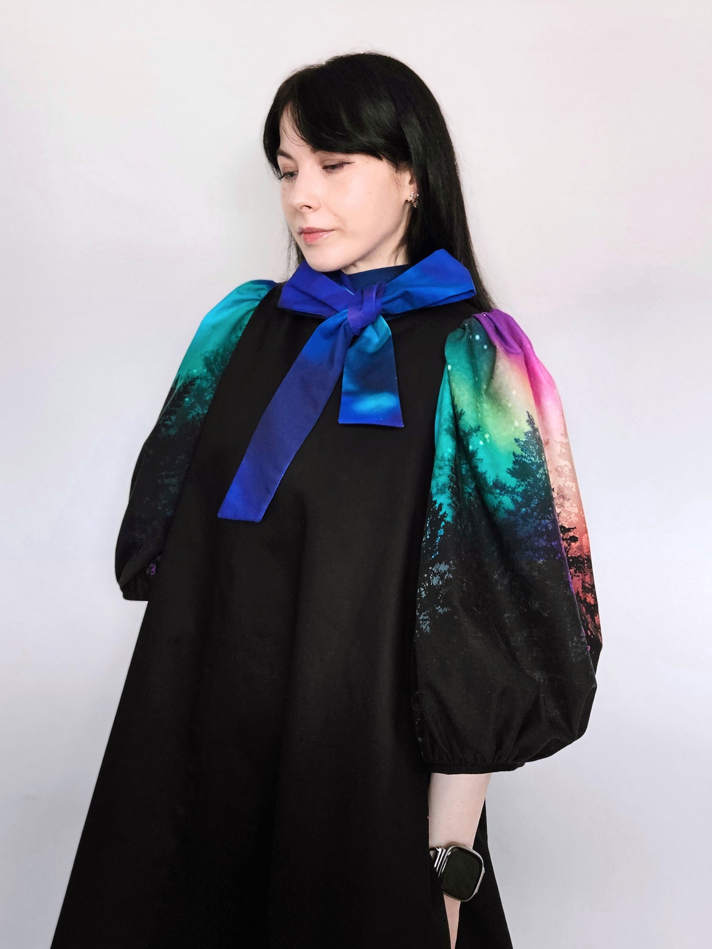 Woman wearing a black dress with colourful aurora print sleeves and a blue bow against a white background