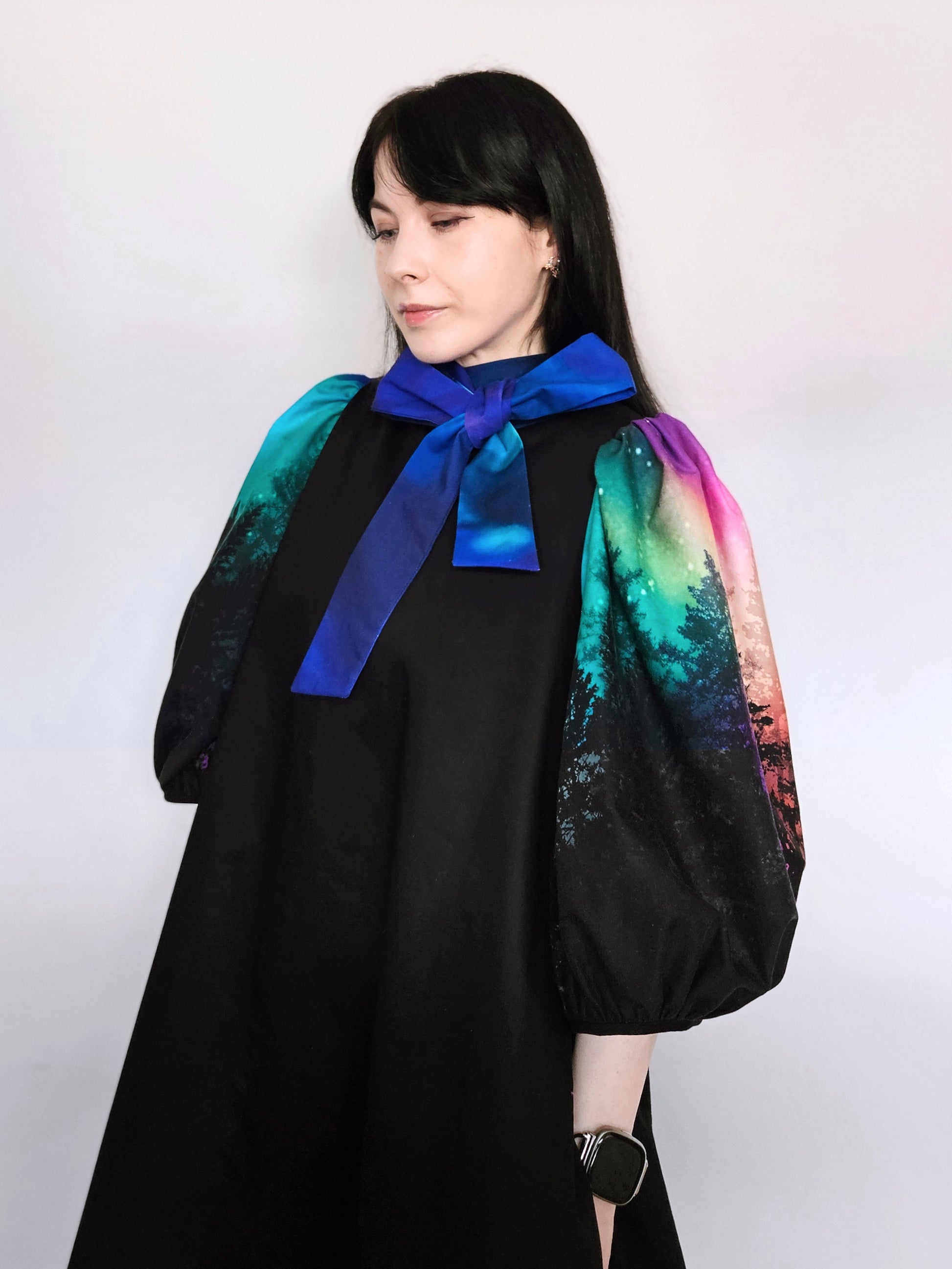 Woman wearing a black dress with colourful aurora print sleeves and a blue bow against a white background