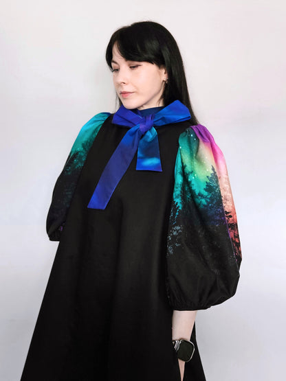Woman wearing a black dress with colourful aurora print sleeves and a blue bow against a white background