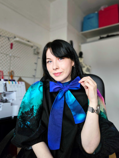 Woman wearing a black dress with colourful aurora print sleeves and a blue bow against a white background