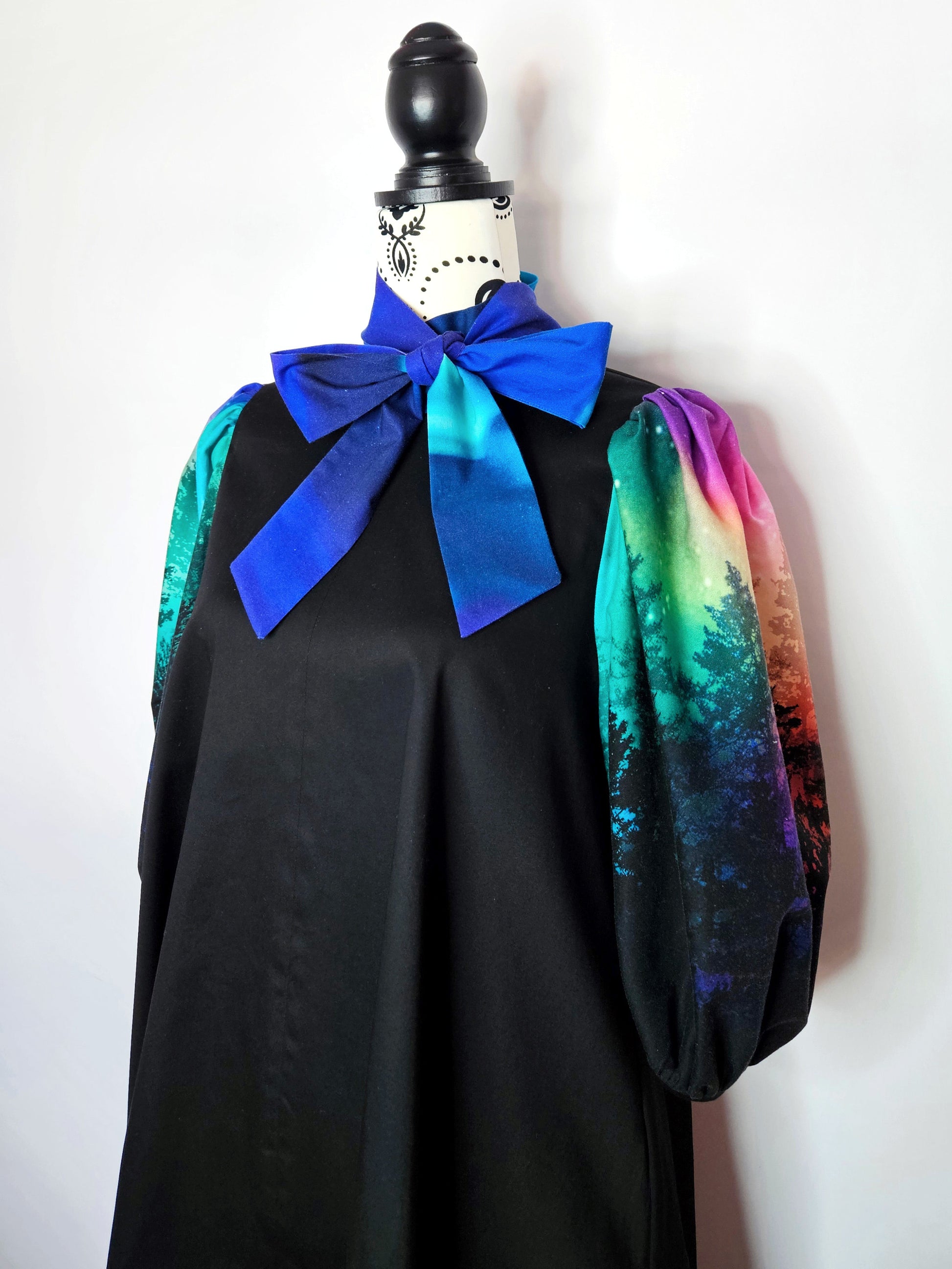 Black dress with colourful aurora print sleeves and blue bow on a mannequin against a white background