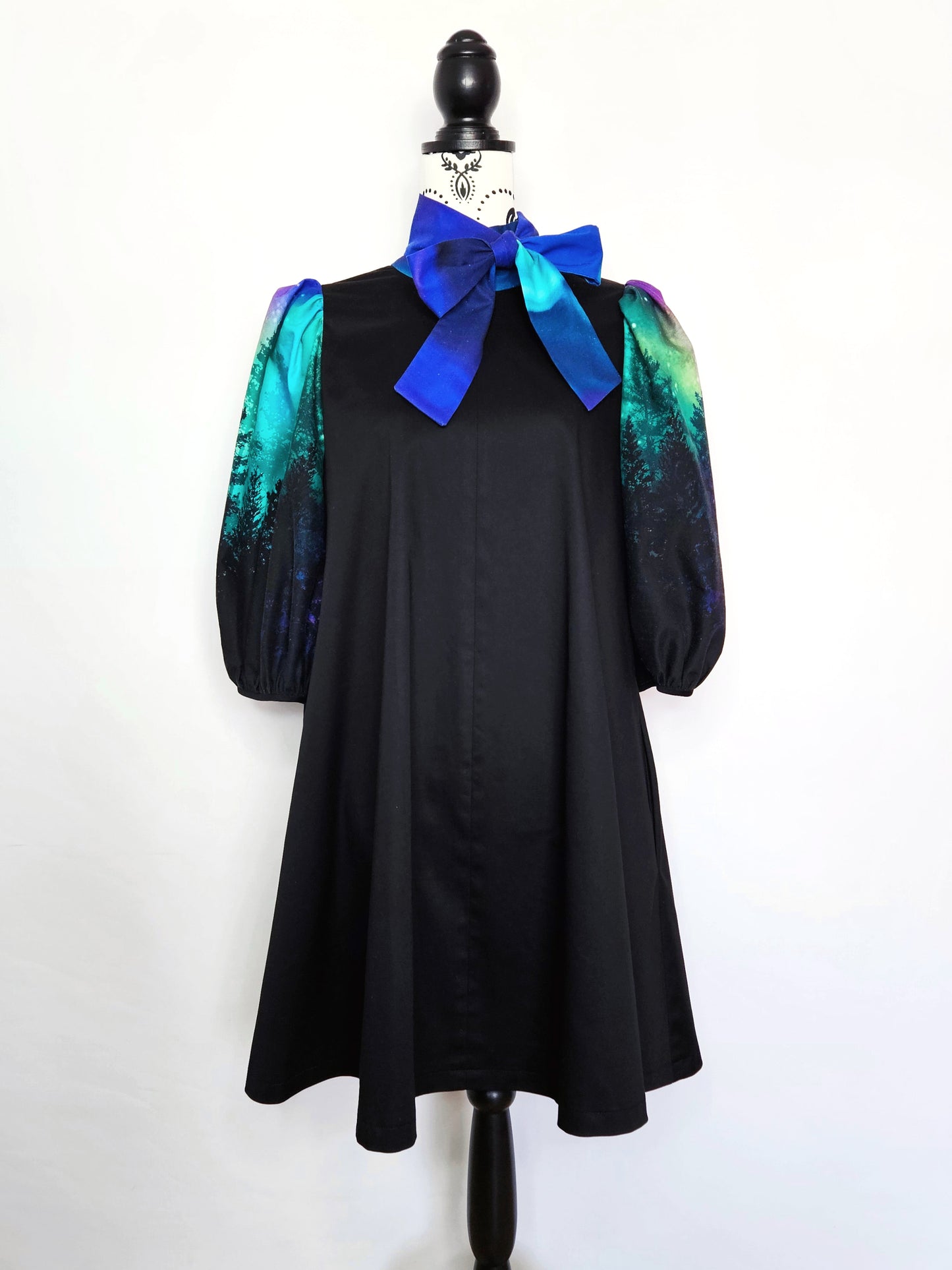 Black dress with colourful aurora print sleeves and blue bow on a mannequin against a white background