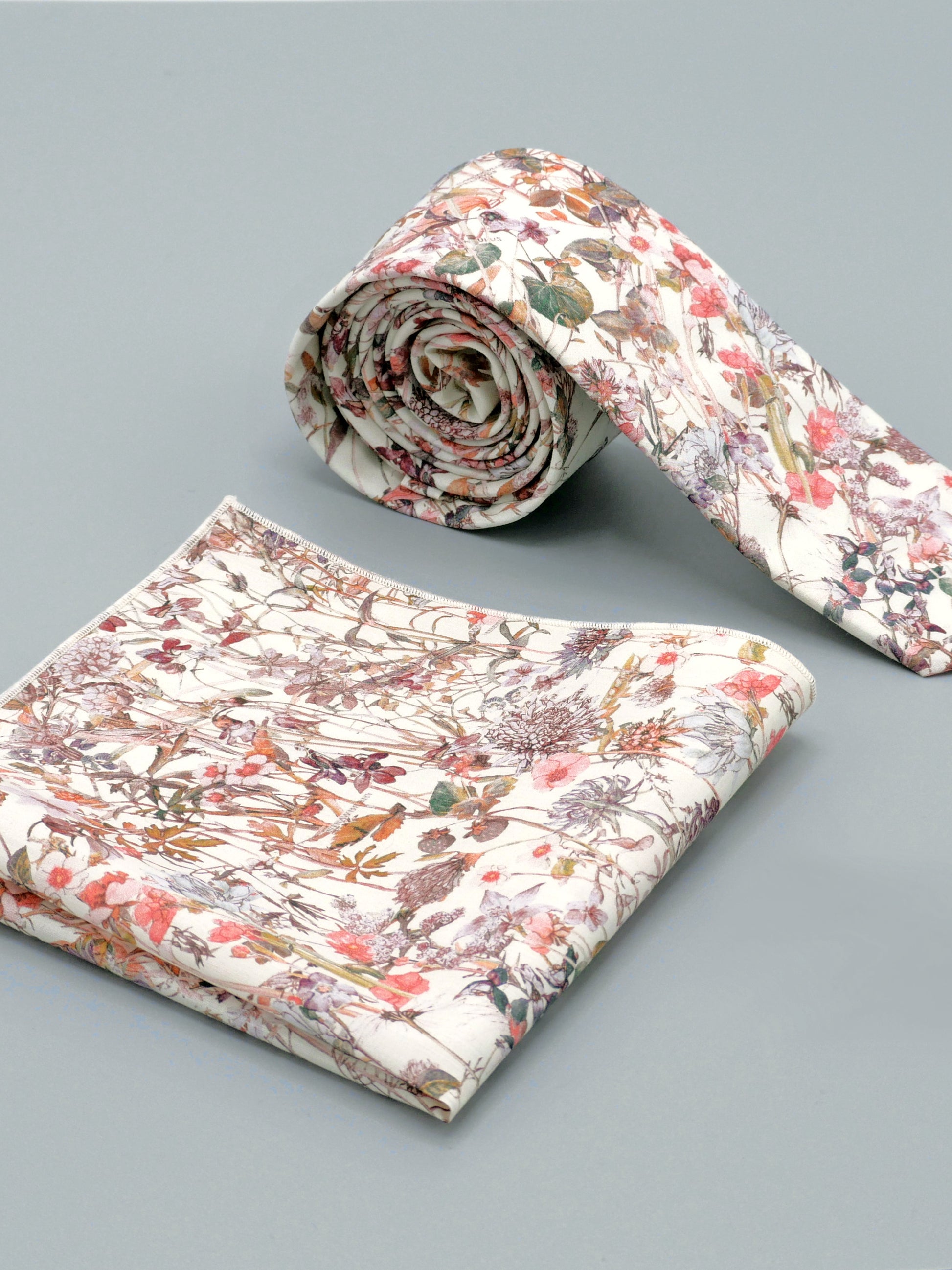 Floral patterned tie in a soft peach pink wildflower print and a matching pocket square placed on a light grey background