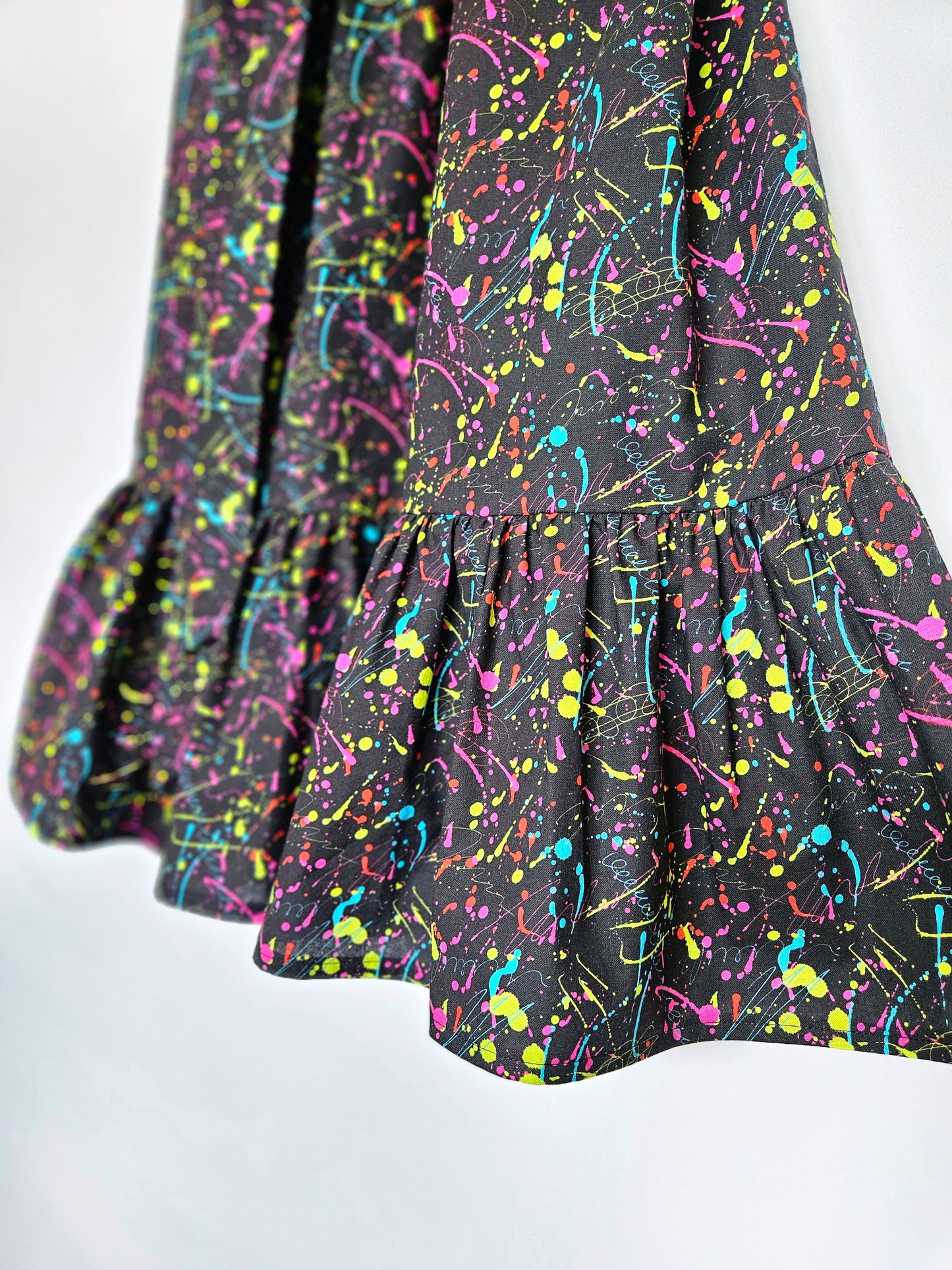 A close-up of a black fabric garment with a vibrant neon paint splatter pattern in pink, blue, yellow, and green, featuring a ruffled hem.