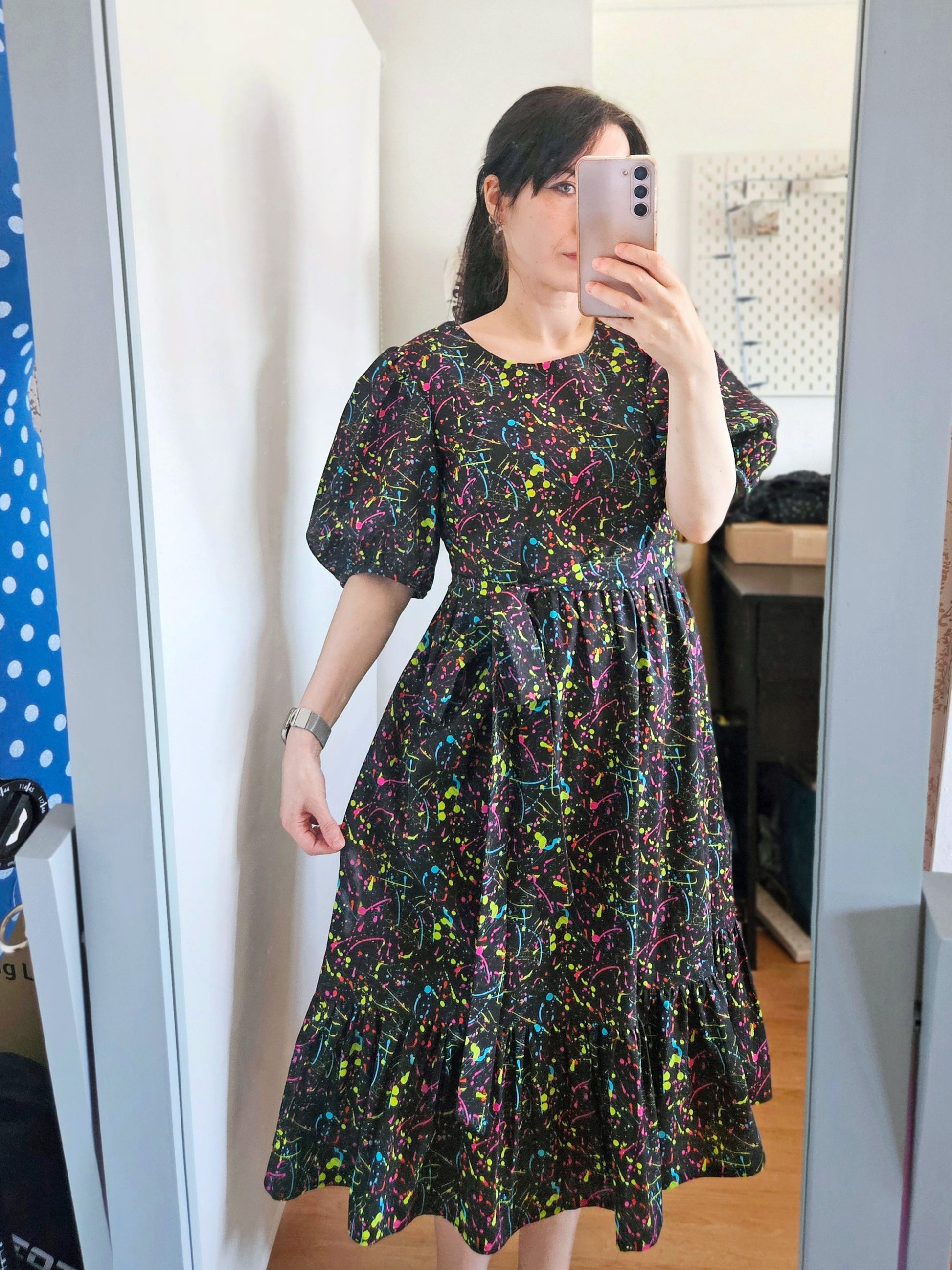 A woman takes a mirror selfie wearing a short-sleeved black dress with a vibrant multicolour paint splatter pattern on a black background.