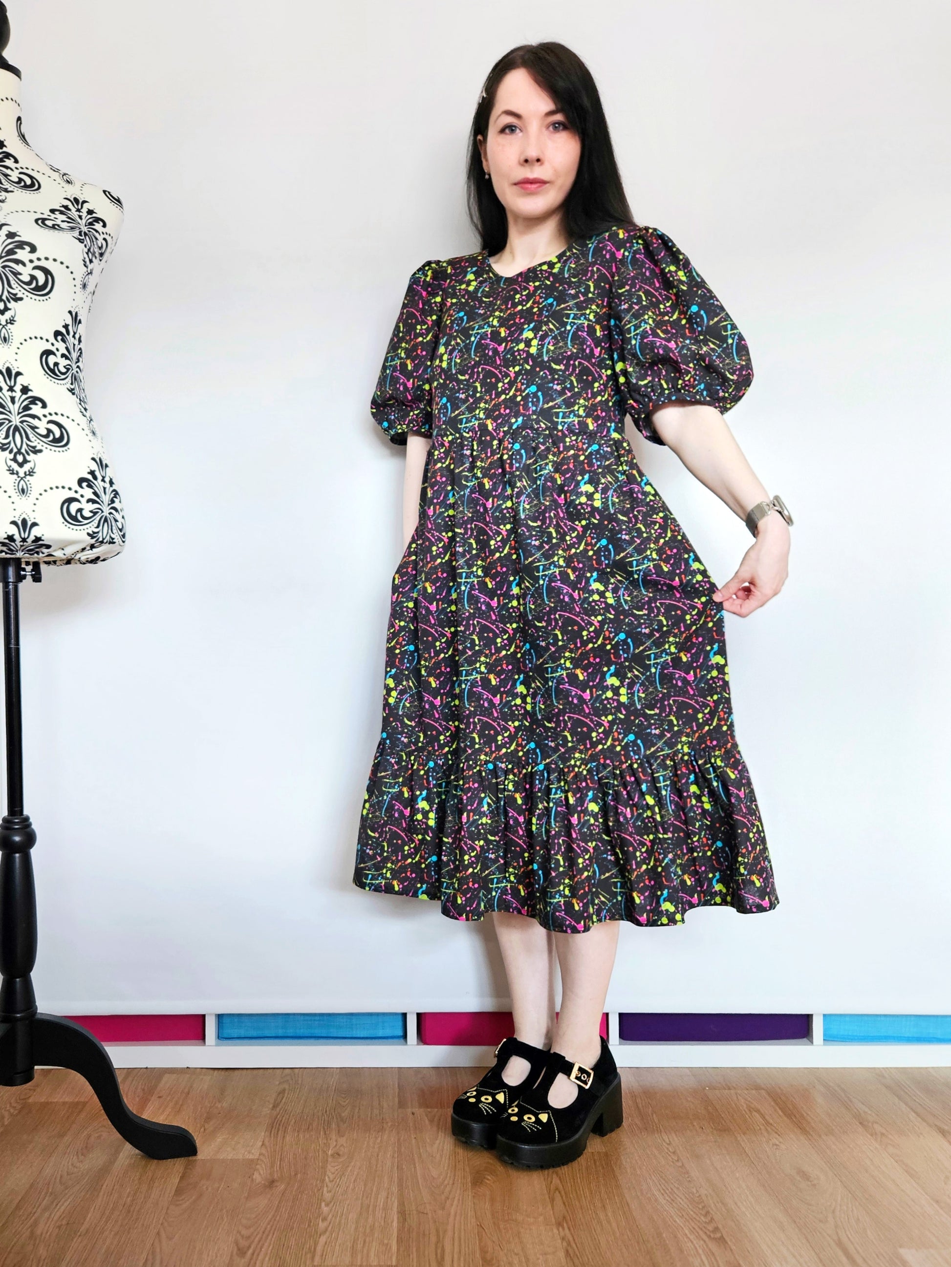 A woman wearing a short puff-sleeve midi dress with a colourful neon paint splatter pattern on a black background, standing in a sewing room with a dress form in the background.