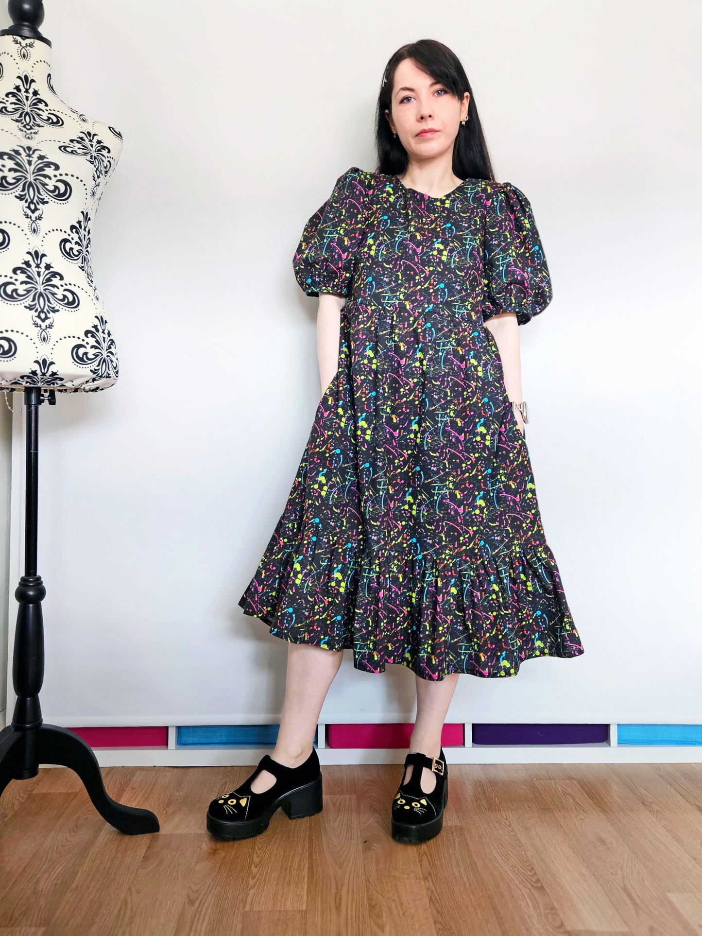 A woman stands indoors wearing a puff-sleeve midi dress with an all-over multicolour paint splatter print on a black background. She is wearing black Mary Jane platform shoes with cat faces.