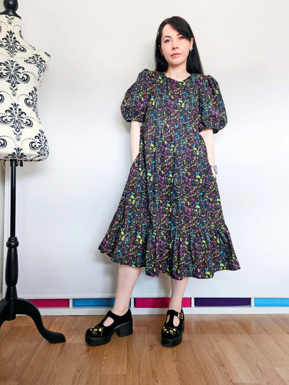 A woman stands indoors wearing a puff-sleeve midi dress with an all-over multicolour paint splatter print on a black background. She is wearing black Mary Jane platform shoes with cat faces.