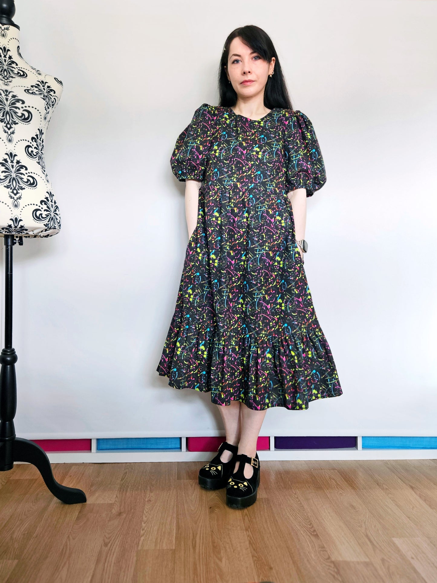 A woman wearing a short puff-sleeve midi dress with a colourful neon paint splatter pattern on a black background, standing in a sewing room with a dress form in the background.