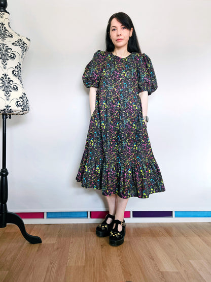 A woman wearing a short puff-sleeve midi dress with a colourful neon paint splatter pattern on a black background, standing in a sewing room with a dress form in the background.
