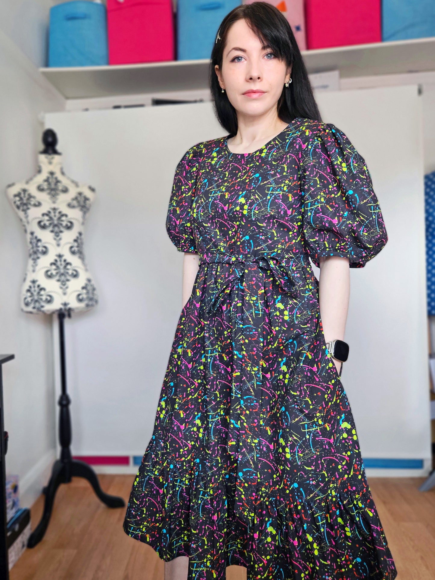 A woman wearing a short puff-sleeve midi dress with a colourful neon paint splatter pattern on a black background, standing in a sewing room with a dress form and shelves of fabric in the background.