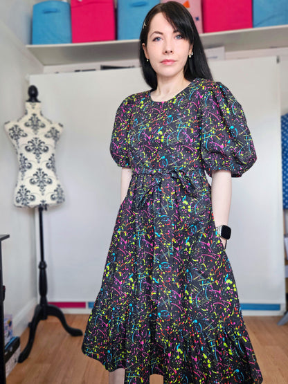 A woman wearing a short puff-sleeve midi dress with a colourful neon paint splatter pattern on a black background, standing in a sewing room with a dress form and shelves of fabric in the background.