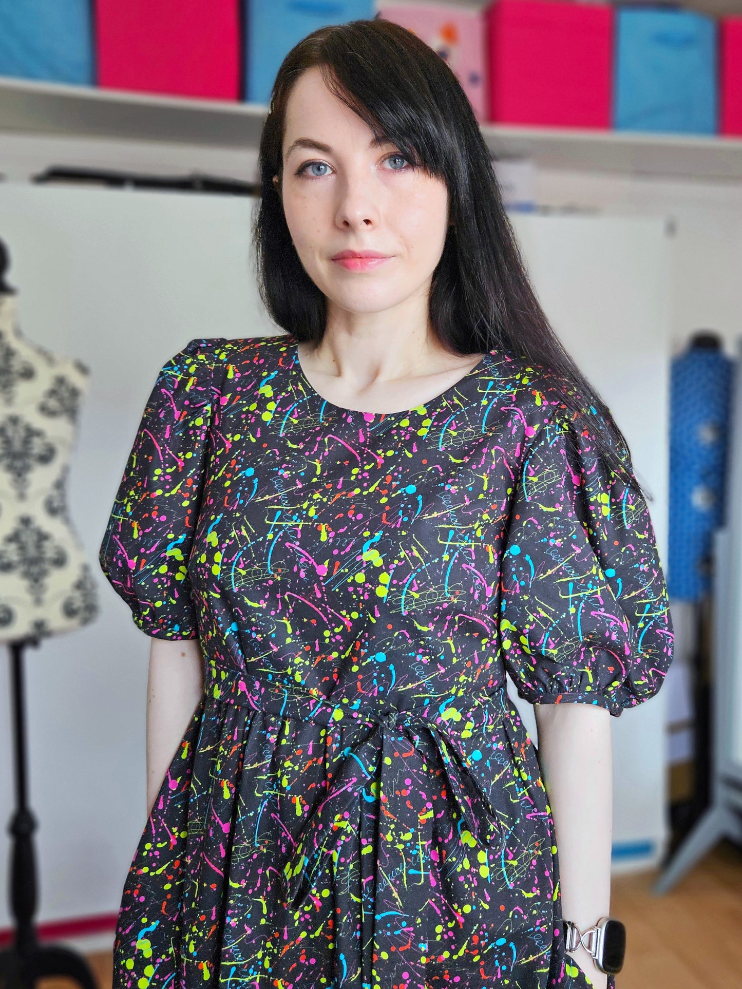 A woman with long dark hair wearing a black puff-sleeve dress with a vibrant multicolour paint splatter pattern, standing in a room with shelves of colourful boxes in the background.