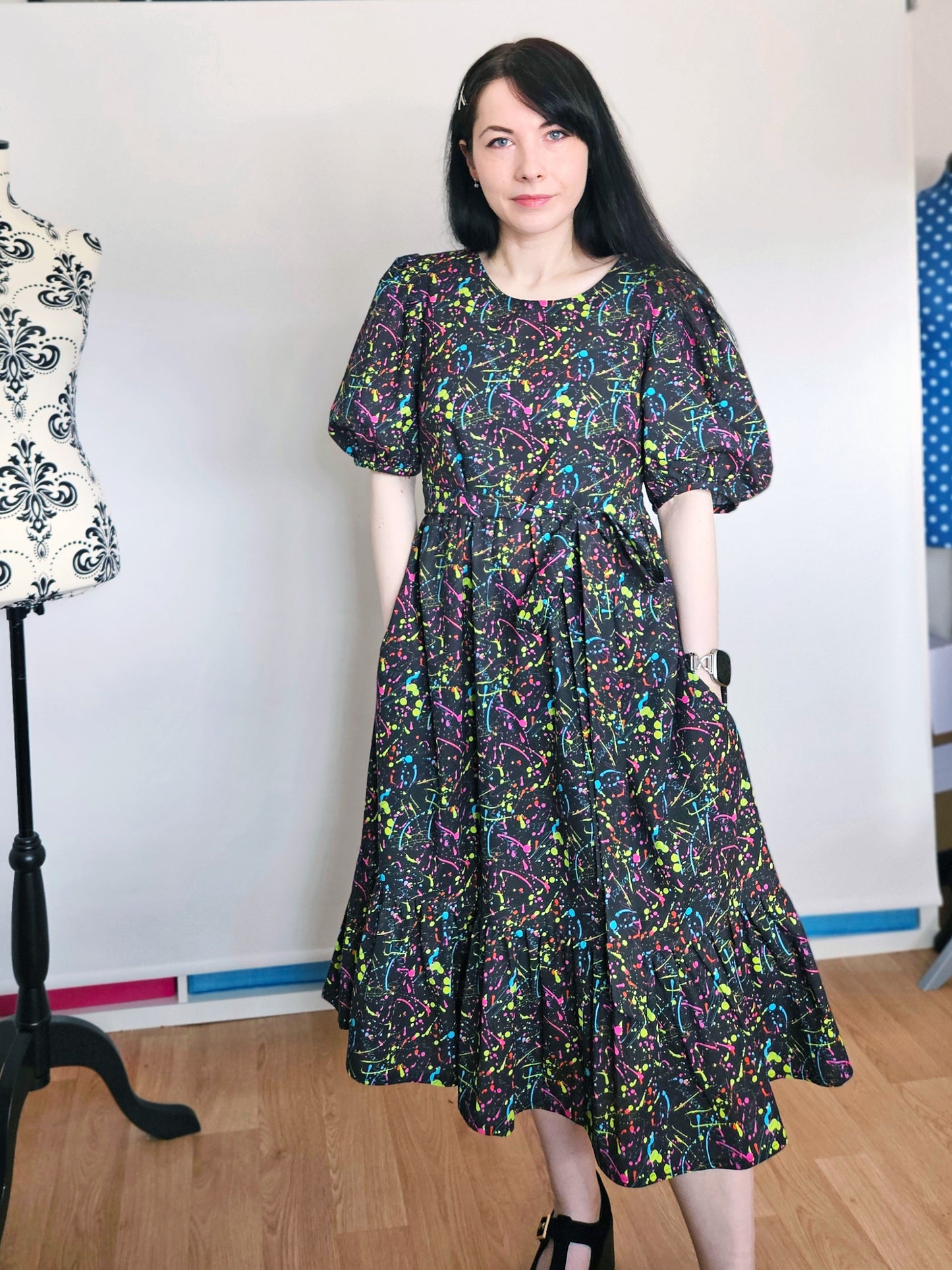 A woman wearing a short-sleeved midi dress with a colorful abstract paint splatter pattern on a black background, standing in a room with a wooden floor and a white wall.