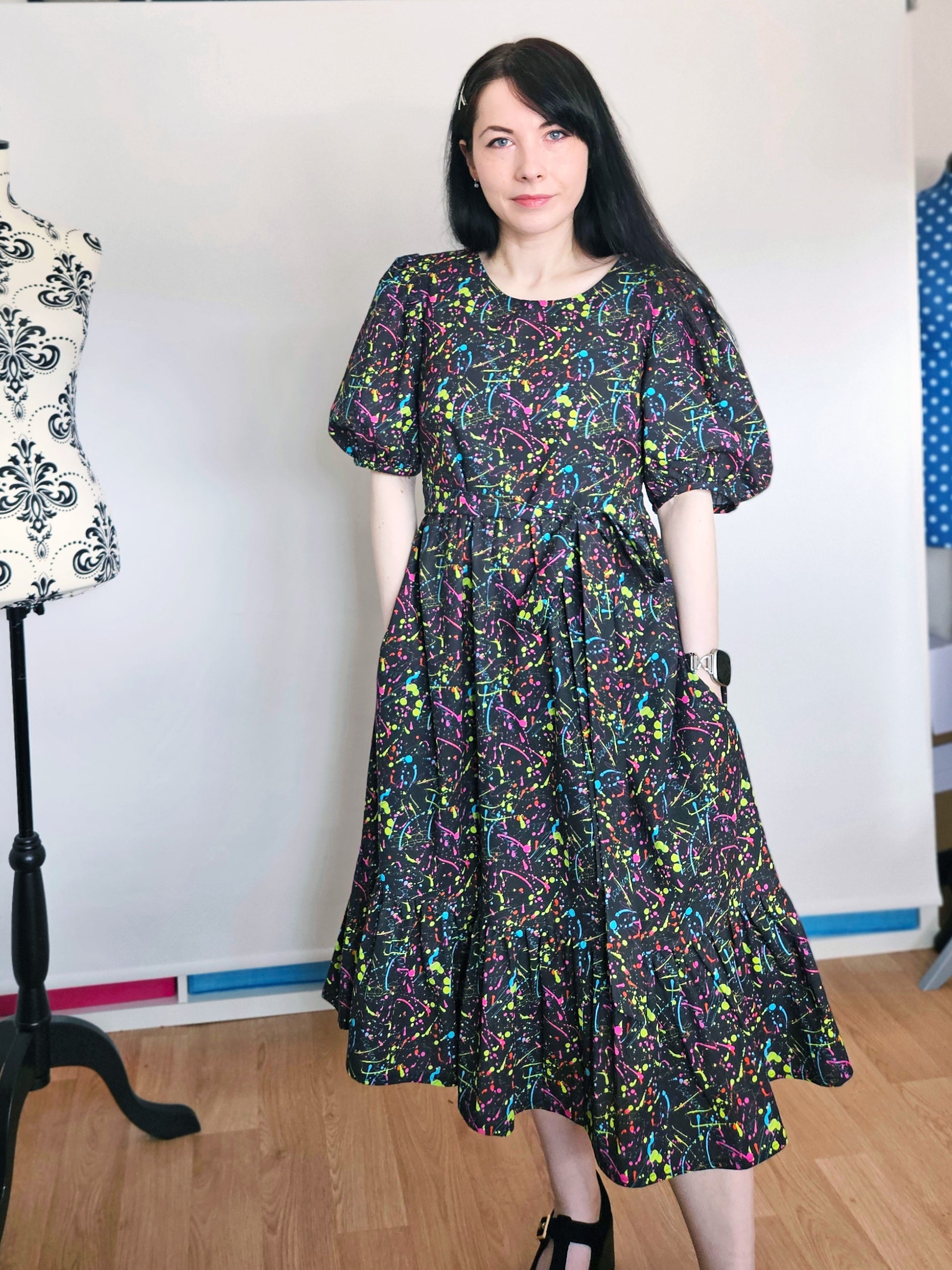 A woman wearing a short-sleeved midi dress with a colorful abstract paint splatter pattern on a black background, standing in a room with a wooden floor and a white wall.