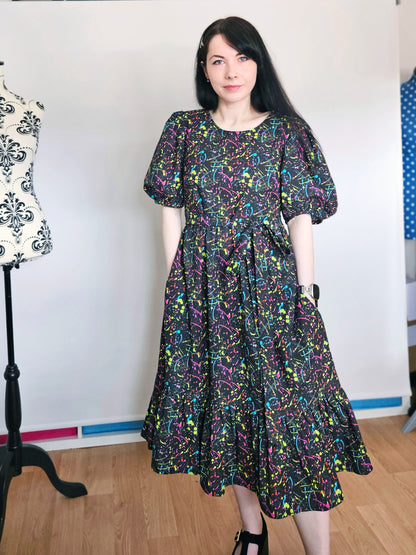 A woman wearing a short-sleeved midi dress with a colorful abstract paint splatter pattern on a black background, standing in a room with a wooden floor and a white wall.