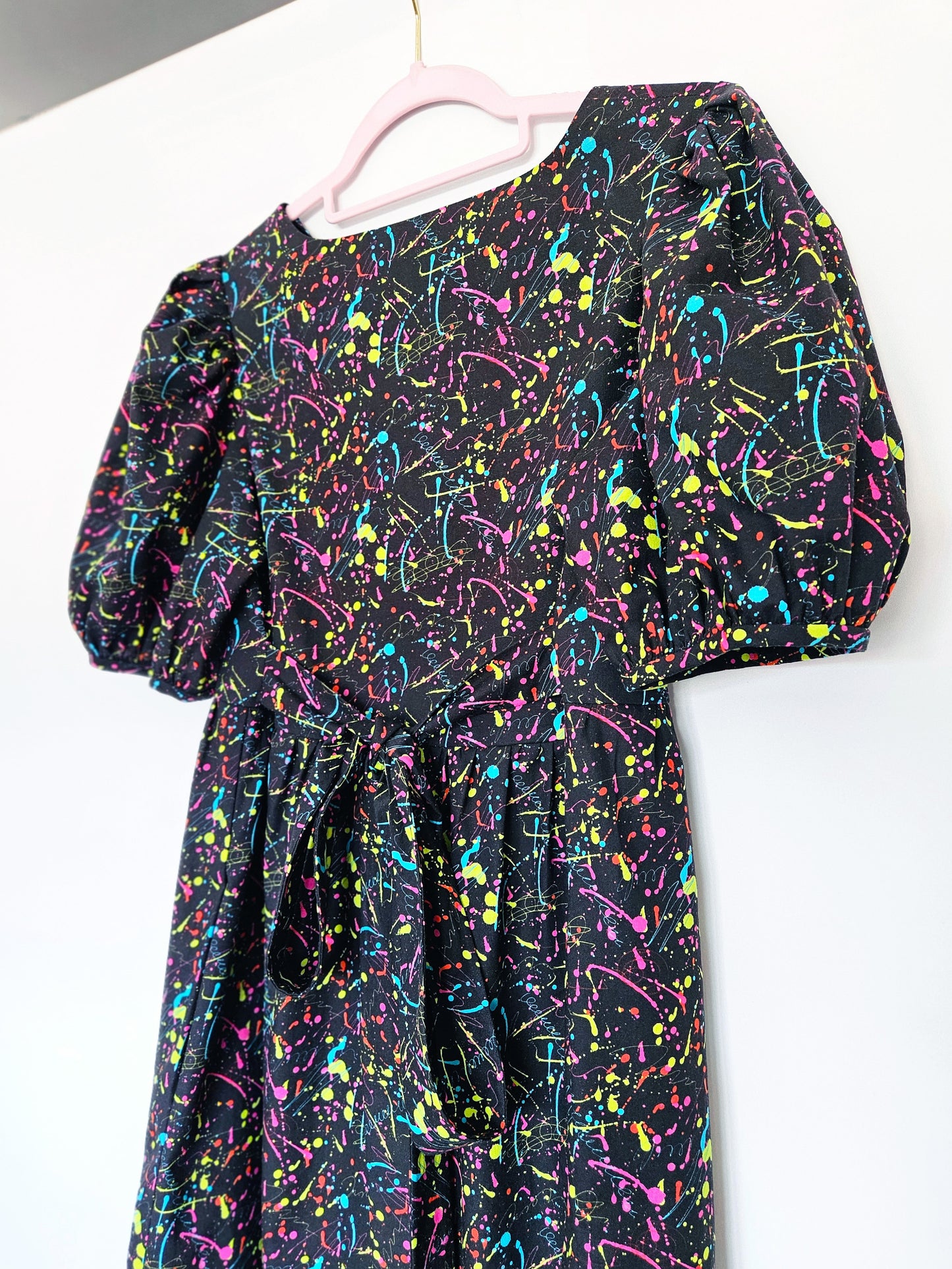 A black dress with a vibrant multicolour paint splatter pattern, featuring puff sleeves and a tie waist, hanging on a light pink hanger against a plain white wall.