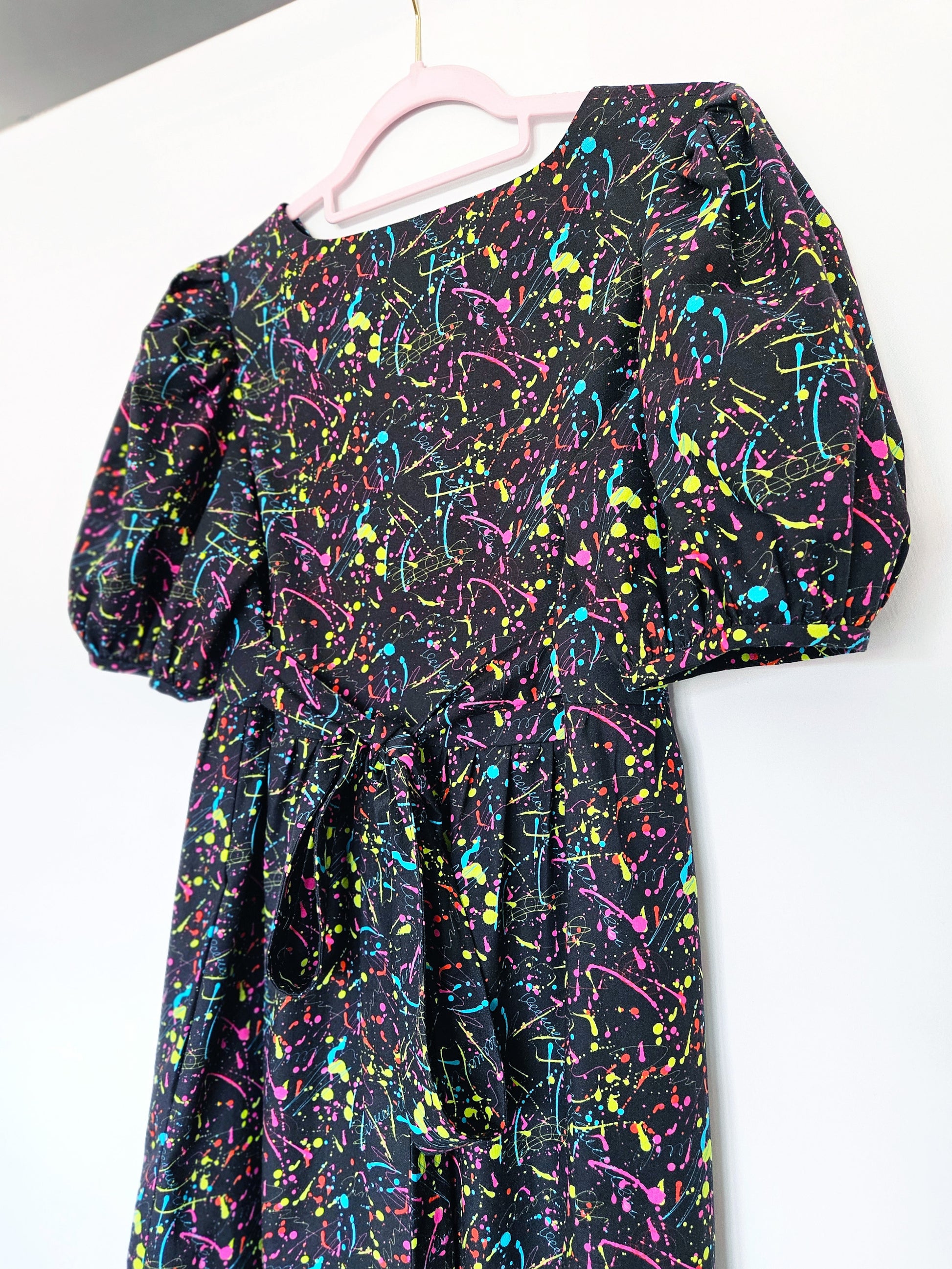 A black dress with a vibrant multicolour paint splatter pattern, featuring puff sleeves and a tie waist, hanging on a light pink hanger against a plain white wall.
