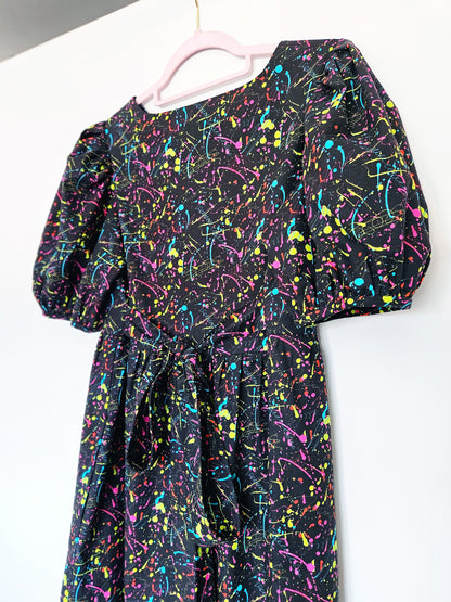 A black dress with a vibrant multicolour paint splatter pattern, featuring puff sleeves and a tie waist, hanging on a light pink hanger against a plain white wall.