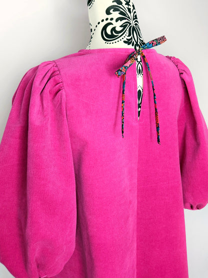 The back of a bright pink dress with puff sleeves on a mannequin against a white wall, showing a floral tie.