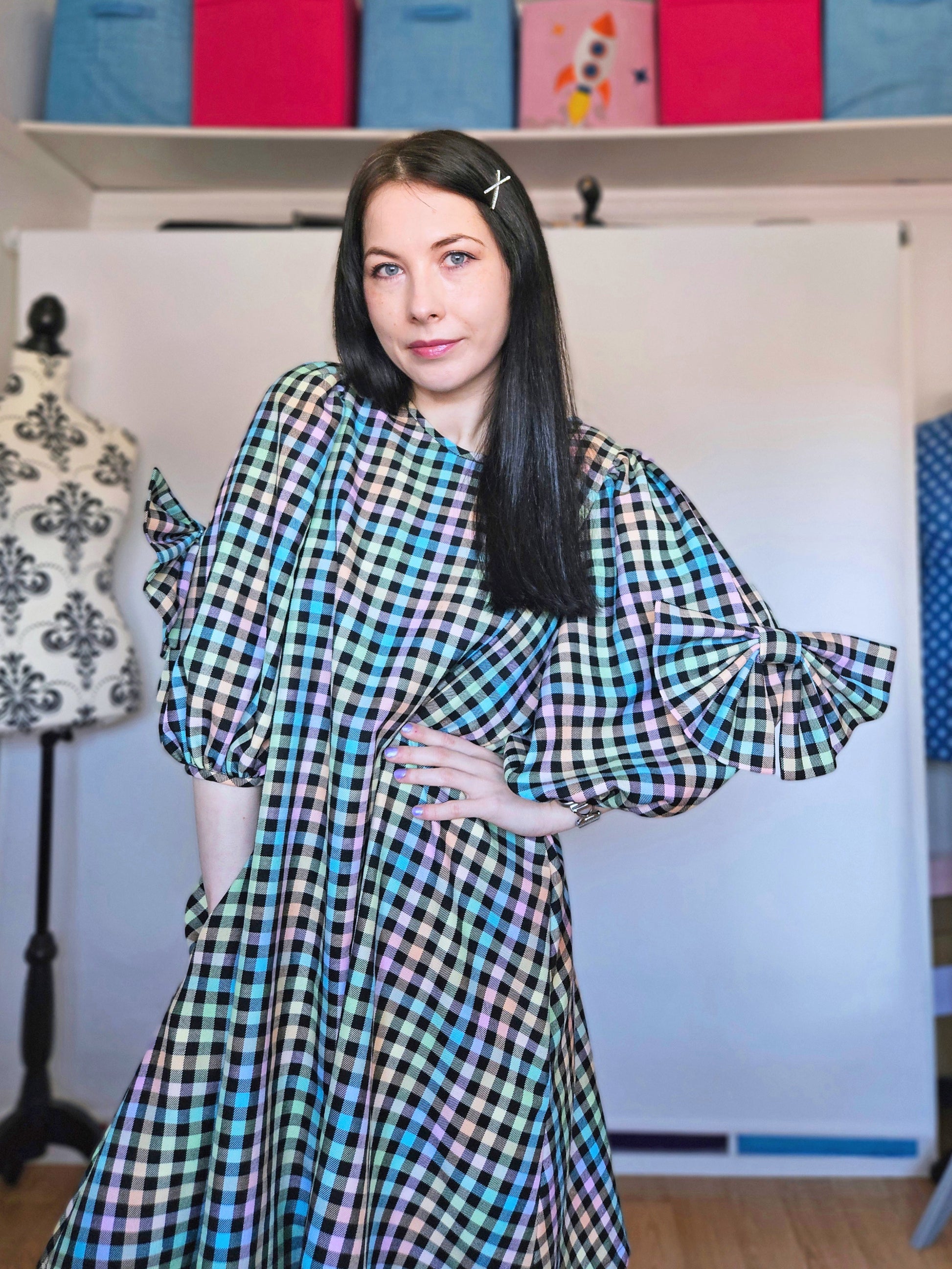 A woman with long dark hair wearing a pastel rainbow tartan dress with voluminous sleeves, standing in front of a white studio backdrop with sewing supplies visible in the background.
