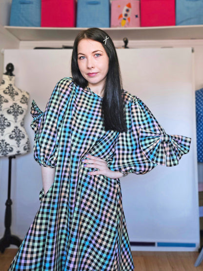 A woman with long dark hair wearing a pastel rainbow tartan dress with voluminous sleeves, standing in front of a white studio backdrop with sewing supplies visible in the background.