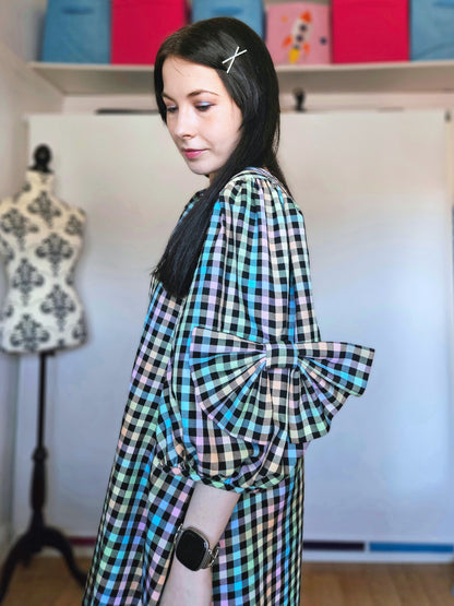 A woman wearing a pastel rainbow checkered dress with oversized puff sleeves, standing in a room with a mannequin and colourful boxes in the background.