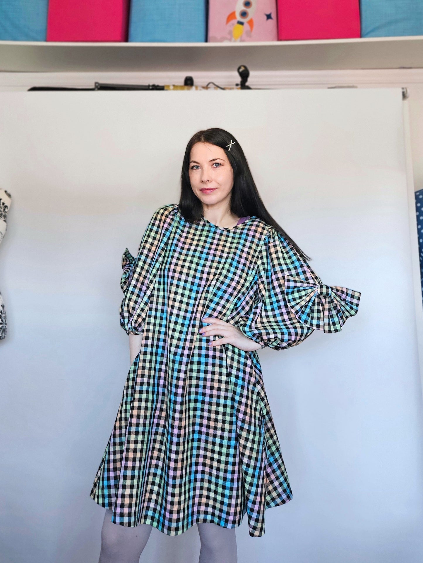 A woman modelling a loose-fitting, knee-length plaid dress with voluminous balloon sleeves and oversized bows. The dress features a black, blue, green and pink pastel rainbow tartan pattern.