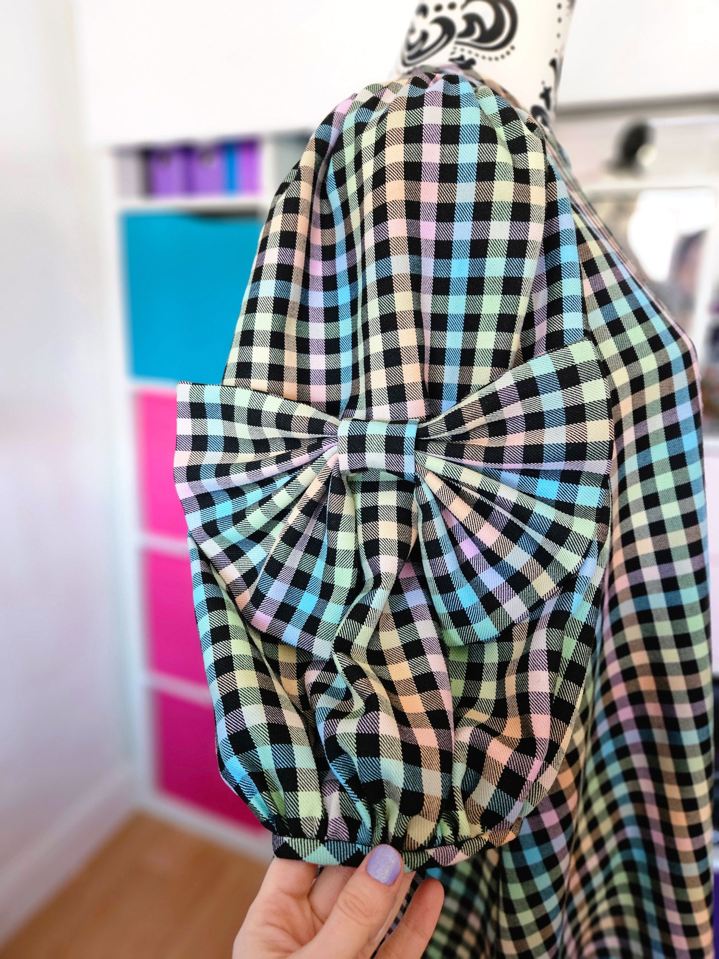 A close-up of a black, white, blue, green, and pink tartan dress with a large bow attached to the sleeve, held by a hand in front of a colourful storage cabinet.