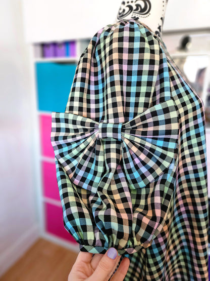 A close-up of a black, white, blue, green, and pink tartan dress with a large bow attached to the sleeve, held by a hand in front of a colourful storage cabinet.