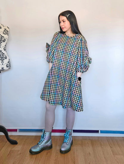 A woman stands indoors wearing a loose, knee-length plaid dress with puff sleeves, paired with beige tights and glittery pastel Dr Martens boots with blue laces. The dress has a multicolour pastel tartan pattern with blue, green, pink yellow, and black.